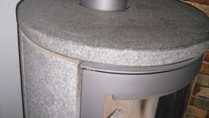 Ein runder Specksteinofen steht in einem modernen Raum, mit einem Kaminrohr oben und einer gläsernen Front, die Holz zeigt.