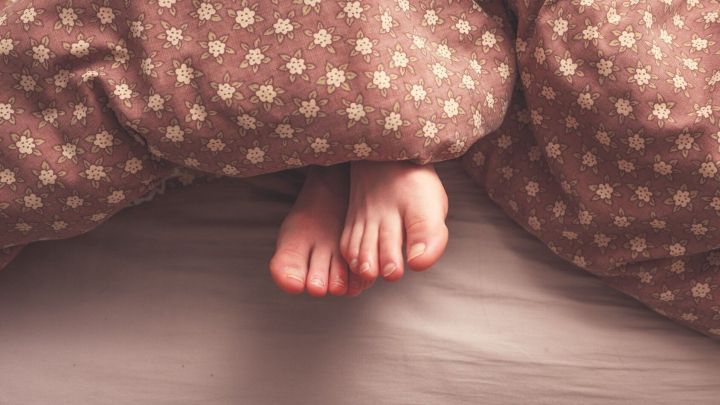 F&uuml;&szlig;e ragen unter einer gemusterten Decke hervor, was M&uuml;digkeit und den Wunsch nach mehr Schlaf signalisiert.