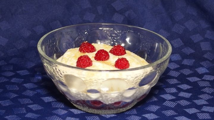 Ein Glas mit Himbeeren und einer cremigen Schichtung steht auf einem blauen Tischcloth und l&auml;dt zum Genie&szlig;en ein.