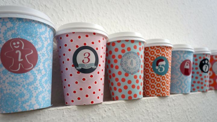 Bunte Coffee-to-go-Becher stehen an einer Wand, dekoriert mit Zahlen f&uuml;r einen Adventskalender.