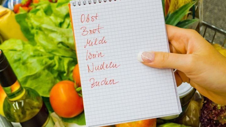 Eine Hand h&auml;lt einen Notizblock mit einer Einkaufsliste. Im Hintergrund sind frische Lebensmittel wie Obst und Gem&uuml;se sichtbar.