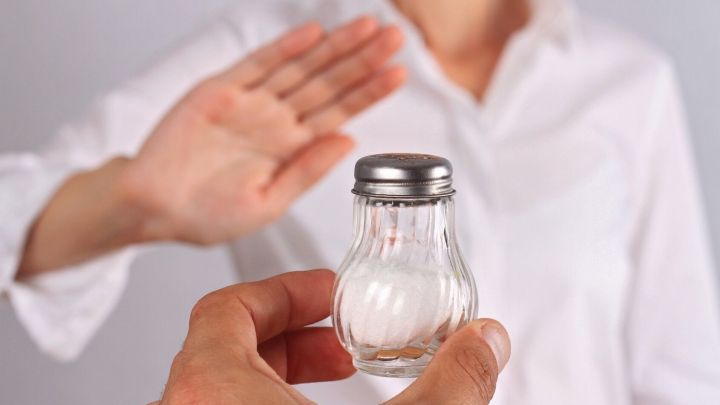 Eine Hand h&auml;lt ein Salzstreuer, w&auml;hrend eine Person mit einer wei&szlig;en Bluse abwehrend die Hand hebt, um Salz abzulehnen.