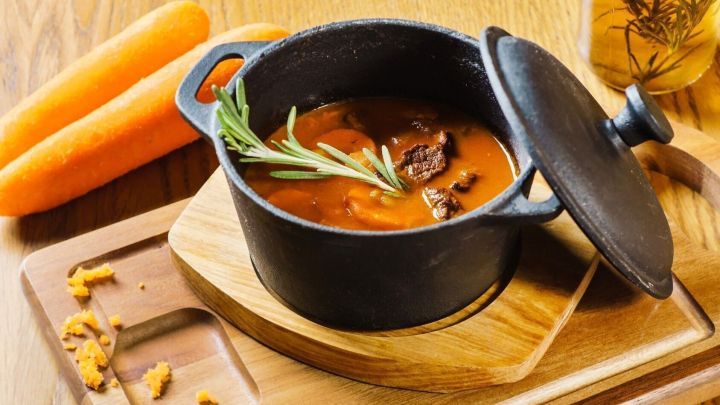 Ein schwerer Karottentopf mit einer duftenden Suppe ist auf einem Holzbrett platziert, umgeben von frischen Karotten.