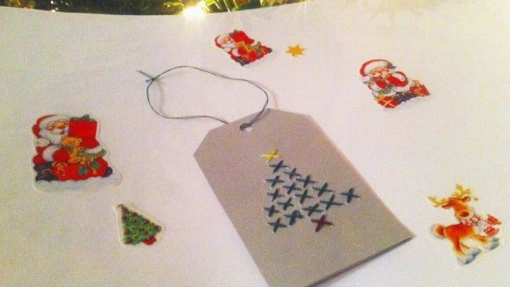 Eine bestickte Geschenkanh&auml;nger liegt auf einem Tisch, umgeben von weihnachtlichen Aufklebern und sanftem Licht.