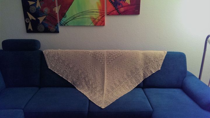 Ein handgestricktes f&uuml;nf-eckiges Dreieckstuch liegt elegant &uuml;ber der blauen Couch in einem hellen Wohnzimmer.