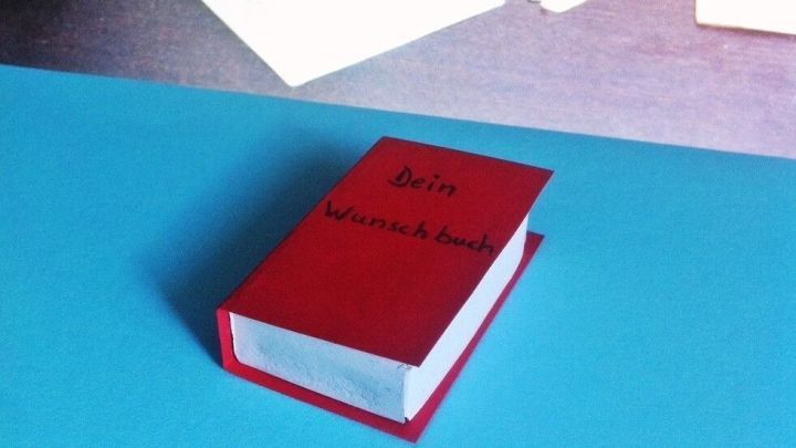 Ein rotes Mini-Buch liegt auf einem blauen Untergrund, umgeben von alten Papierunterlagen und Rosen aus Papier, ideal als Geldgeschenk.