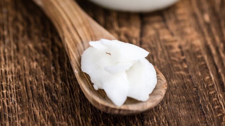 Ein Glas Kokos&ouml;l steht auf einem Holzuntergrund, w&auml;hrend ein L&ouml;ffel mit einem Klecks des &Ouml;ls daneben liegt.