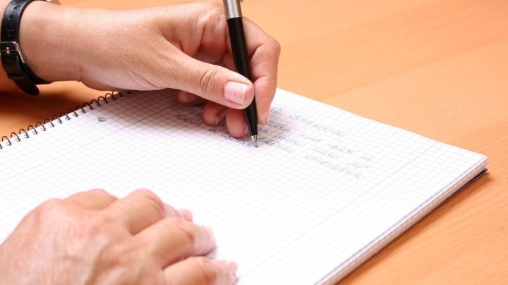 Eine Hand h&auml;lt einen Stift und schreibt auf ein Notizblatt, w&auml;hrend der Tisch eine warme Holzoberfl&auml;che zeigt.