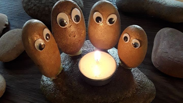 F&uuml;nf mit Wackelaugen versehene Steine sitzen um eine brennende Kerze auf einem Tisch, teilen einen gem&uuml;tlichen Moment.