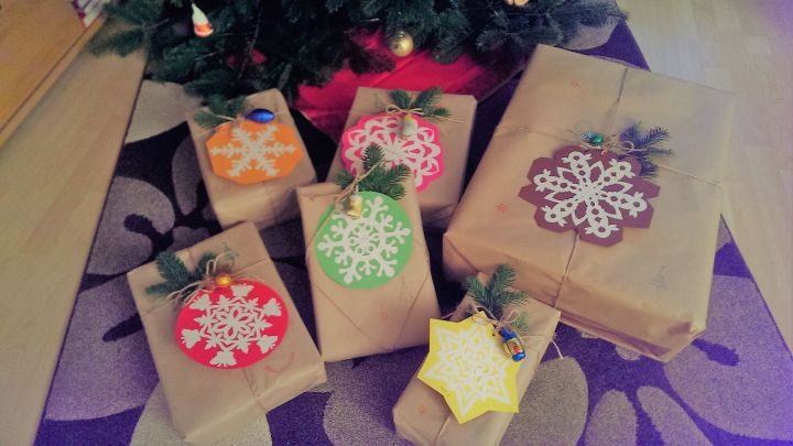 Geschenkp&auml;ckchen aus Kraftpapier liegen um einen Weihnachtsbaum, dekoriert mit bunten Anh&auml;ngern und nat&uuml;rlichem Gr&uuml;n.