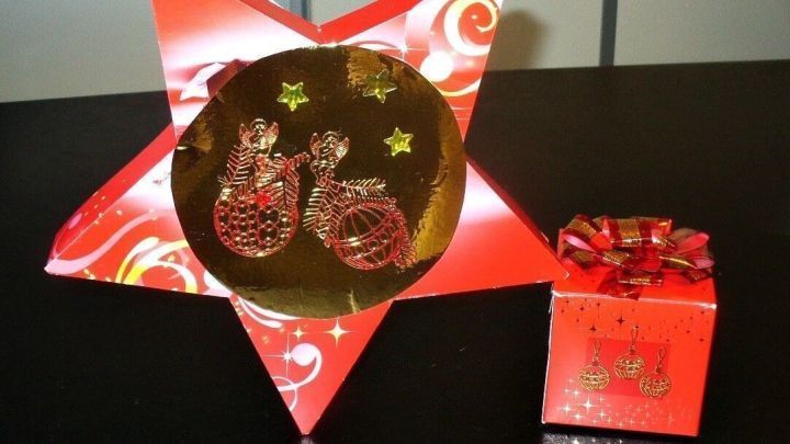 Eine gro&szlig;e, rote Stern-Pralinengeschenkbox steht im Vordergrund, neben einer kleinen, festlichen Geschenkbox auf einem Tisch.