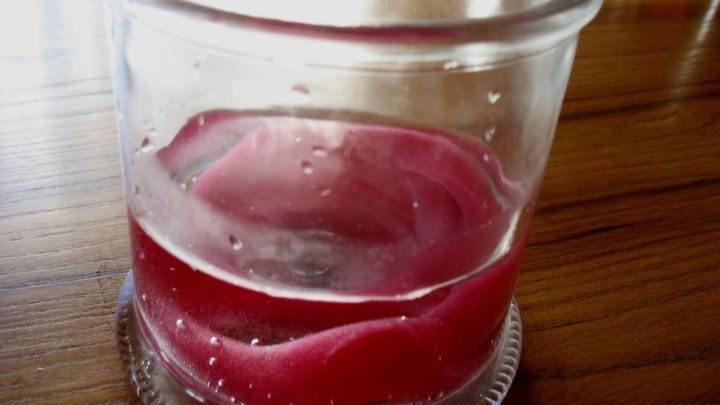 Ein durchscheinendes Glasgef&auml;&szlig; steht auf einem Holztisch, gef&uuml;llt mit geschmolzenem Wachs in r&ouml;tlicher Farbe.