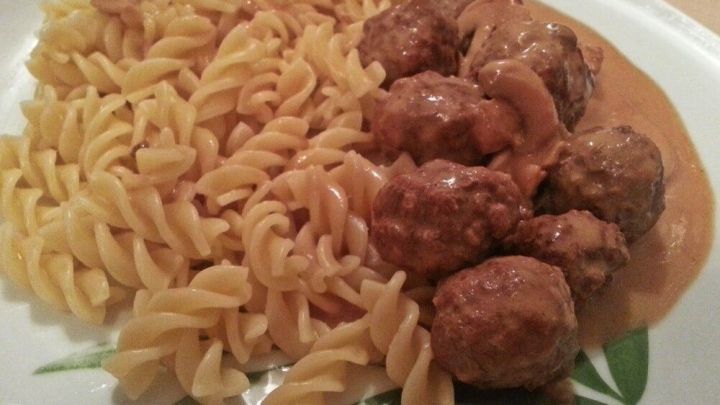 Saftige K&ouml;ttbullar in Rahmsauce liegen neben spiralf&ouml;rmiger Pasta auf einem Teller, umgeben von einem schlichten Tisch.