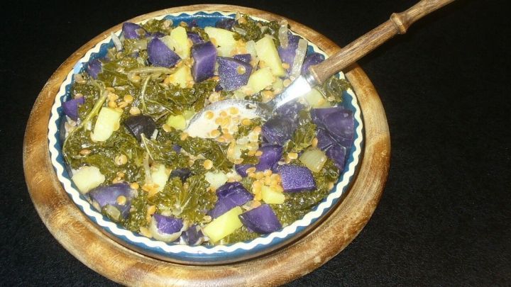 Ein Sch&uuml;ssel Gr&uuml;nkohl mit roten Linsen und lila Kartoffeln wird mit einem L&ouml;ffel serviert, auf schwarzem Untergrund.