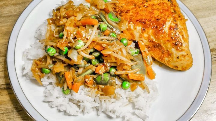 Ein Teller mit Wei&szlig;reis, belegt mit buntem asiatischem Gem&uuml;se und gebratenem H&auml;hnchen, serviert auf einem Tisch.