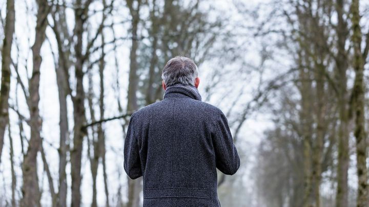 Ein Mann in einem dunklen Mantel geht allein auf einem schmalen Weg durch einen winterlichen Wald, um Winterblues zu bek&auml;mpfen.