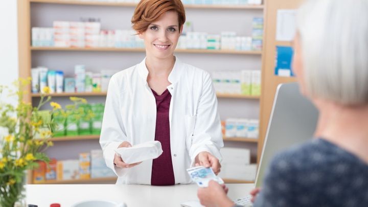 Eine Apothekerin &uuml;berreicht einem &auml;lteren Kunden ein Medikament in einem gesch&auml;ftlichen Umfeld mit Regalen voller Produkte.