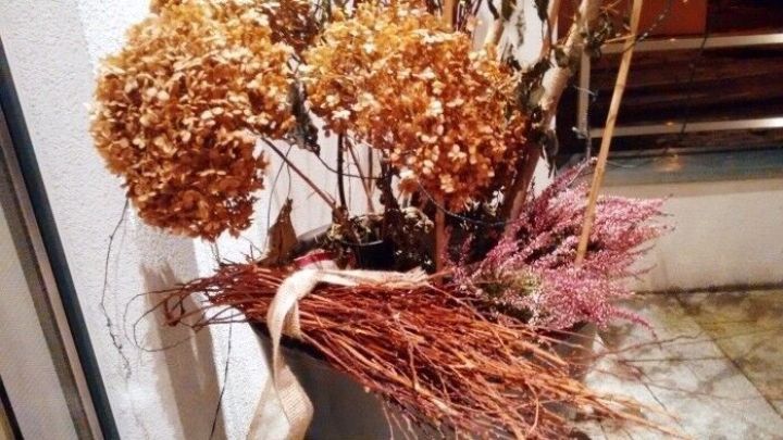 Eine dekorative Grabvase mit getrockneten Blumen und Zweigen steht in einem Blumentopf, umgeben von weiteren Pflanzen.