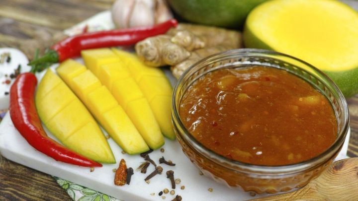 Eine Schale mit chutney steht auf einem Brett, umgeben von Mangos, Chili, Ingwer und Knoblauch, die alle f&uuml;r pikante Saucen verwendet werden.