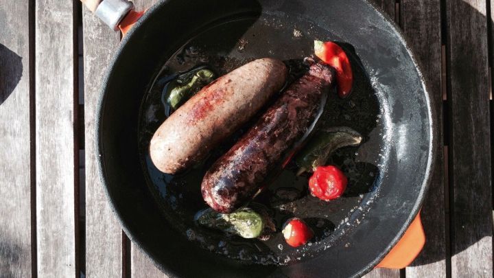 Blut- und Leberw&uuml;rste braten in einer Pfanne, umgeben von frischen Gem&uuml;se auf einer rustikalen Holzoberfl&auml;che.