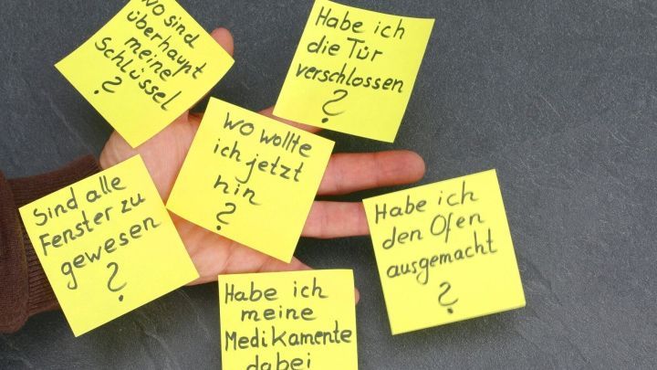 Ein Hand h&auml;lt mehrere gelbe Post-it-Zettel, die Fragen wie &bdquo;Habe ich die T&uuml;r verschlossen?&ldquo; und &bdquo;Wo wollte ich jetzt hin?&ldquo; zeigen.