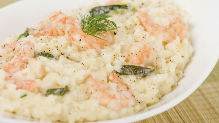 Cremiges Risotto liegt in einer wei&szlig;en Sch&uuml;ssel, garniert mit Scampi und Dill, auf einem hellen Untergrund.
