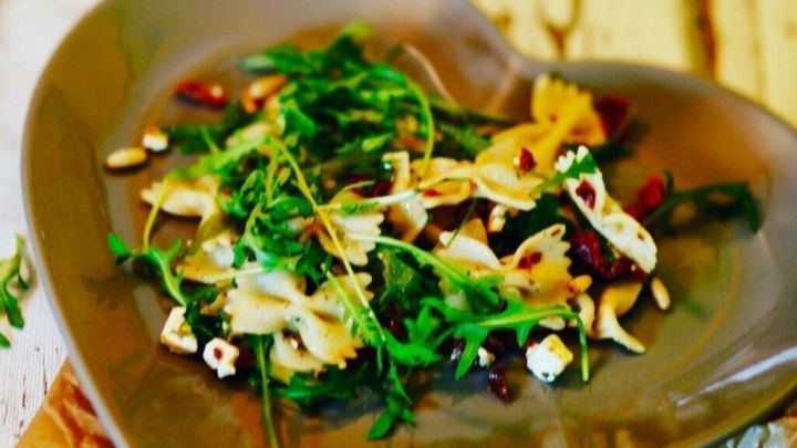 Ein herzf&ouml;rmiger Teller mit Nudelsalat, Rucola und Pinienkernen steht auf einem rustikalen Holztablett, umgeben von dekorativen Salzstreuern.