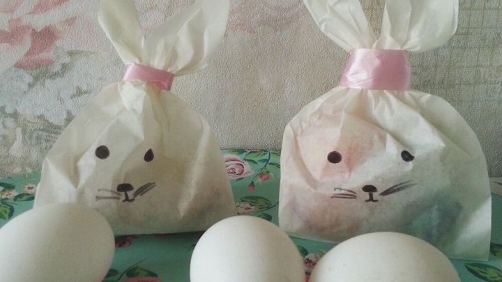 Zwei kleine, dekorative Osterhasen-Geschenkt&uuml;ten stehen auf einem bl&uuml;henden Tisch, daneben liegen drei wei&szlig;e Eier.