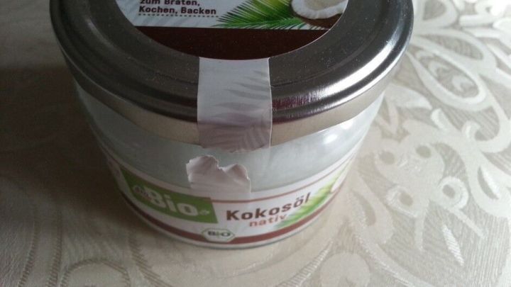 Ein Glas Kokos&ouml;l steht auf einem hellen Tisch. Es zeigt das Etikett mit der Aufschrift "Kokos&ouml;l nativ".