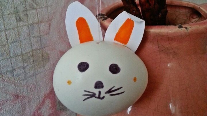 Ein h&auml;ngendes, verziertes Ei mit Hasenohren schaukelt sanft an einem Baumzweig in einer Blumentopfdeko.