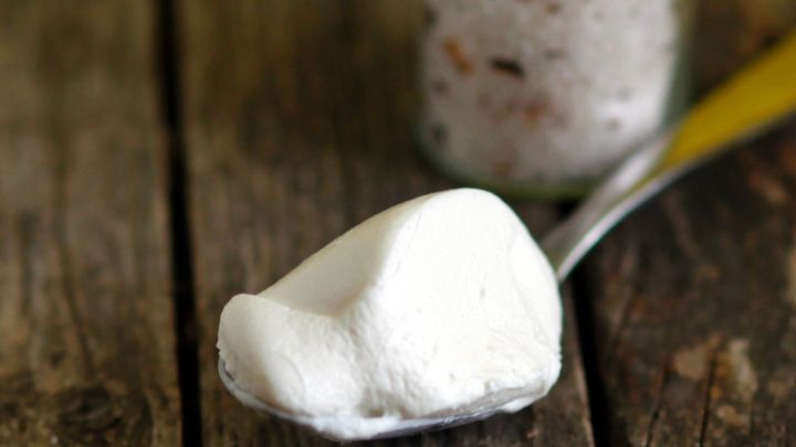 Ein Glas mit Salz steht neben einem L&ouml;ffel, der eine Portion Frischk&auml;se h&auml;lt, auf einem rustikalen Holztisch.