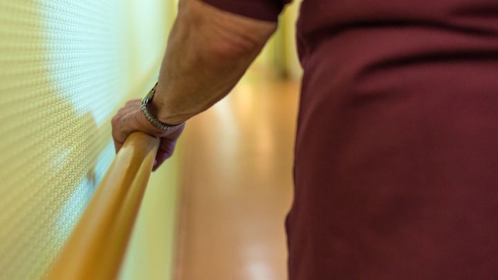 Eine Hand h&auml;lt sich an einem gelben Handlauf, w&auml;hrend eine Person einen Flur entlanggeht, was Sicherheit und Unterst&uuml;tzung symbolisiert.