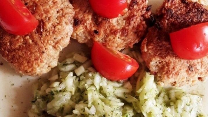 Bratlinge aus Haferflocken liegen auf einem Teller, garniert mit Tomaten, neben einem gr&uuml;nen Gurkensalat.
