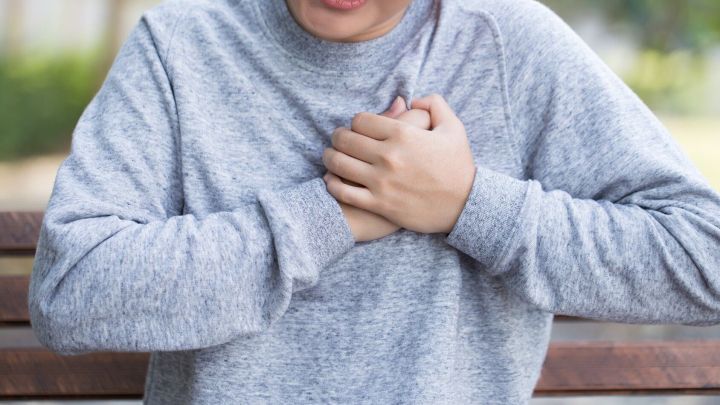 Eine Person fasst sich besorgt an die Brust und zeigt Symptome von Atemnot, w&auml;hrend sie auf einer Bank sitzt.