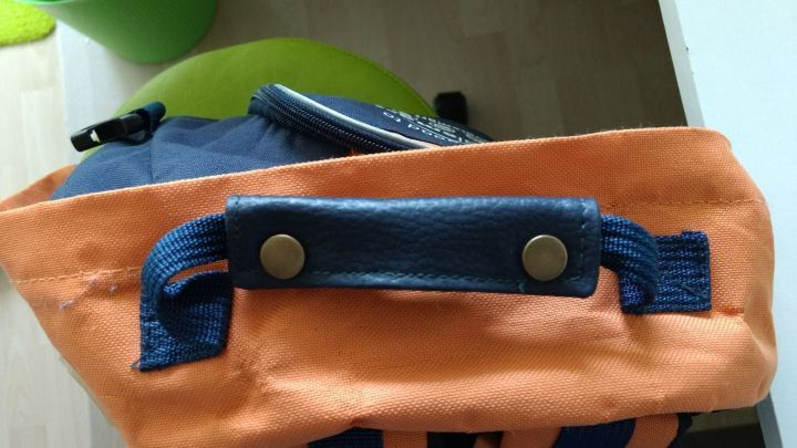 Ein Rucksack mit einem orangefarbenen Griffpolster, das stabil angen&auml;ht ist, liegt auf einem Tisch in einem hellen Raum.