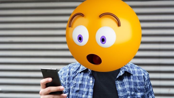 Ein erstaunter Emoji-Kopf sieht auf ein Smartphone, w&auml;hrend er in einem urbanen Umfeld steht, umgeben von einer gestreiften Wand.