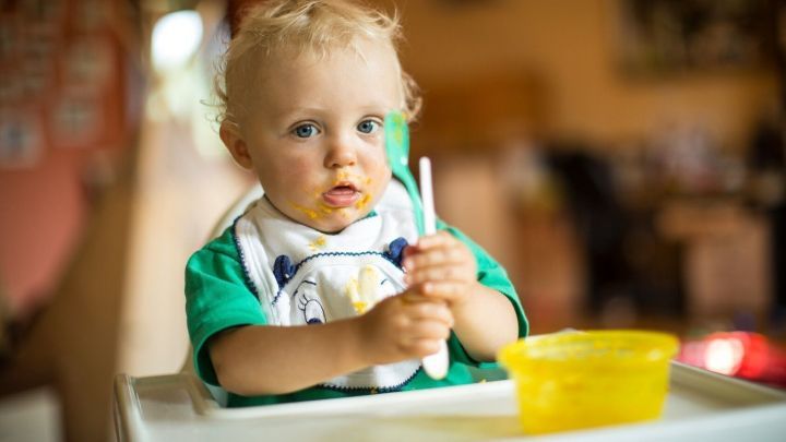 Ein kleines Kind sitzt im Hochstuhl, h&auml;lt einen L&ouml;ffel in der Hand und schaut konzentriert auf eine gelbe Sch&uuml;ssel.