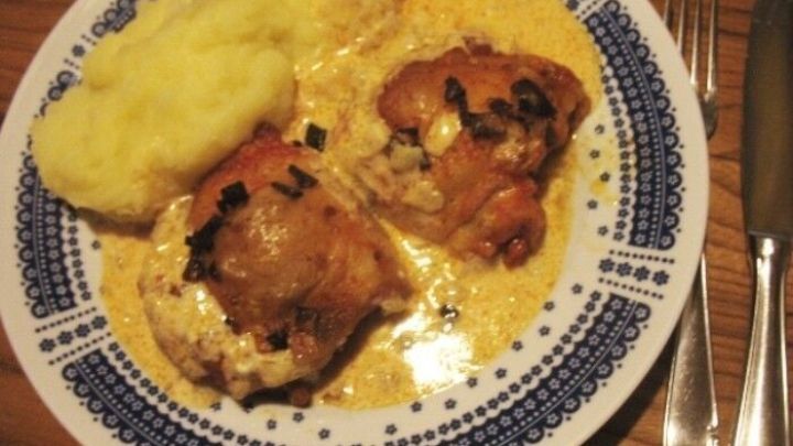 Zarte &uuml;berbackene H&auml;hnchensteaks liegen auf einem Teller mit cremiger Senf-Schmand-Sauce und Beilagen, umgeben von Besteck.