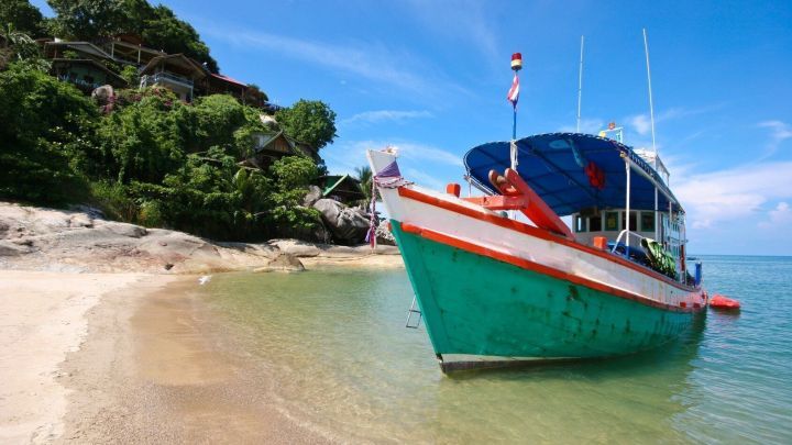 Ein buntes Boot liegt am Strand, w&auml;hrend sanfte Wellen an das Ufer pl&auml;tschern, umgeben von gr&uuml;nem Ufer und blauen Himmel.