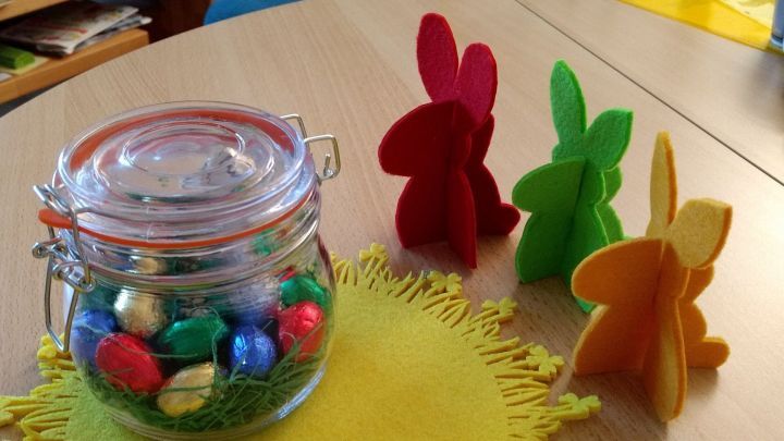 Ein Glas mit bunten Schokoladeneiern steht auf einem Tisch, umgeben von farbigen Papp-Osterhasen in fr&ouml;hlicher Atmosph&auml;re.