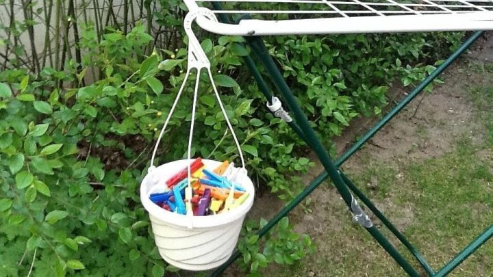 Eine Blumenampel als Klammerk&ouml;rbchen h&auml;ngt an einem W&auml;schegestell und enth&auml;lt bunte W&auml;scheklammern, umgeben von Pflanzen.