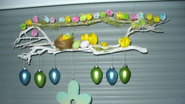 Eine bunte Oster-K&uuml;kenfamilie sowie Eier und eine Holzblume h&auml;ngen aktiv an einem Ast, der festlich dekoriert ist.