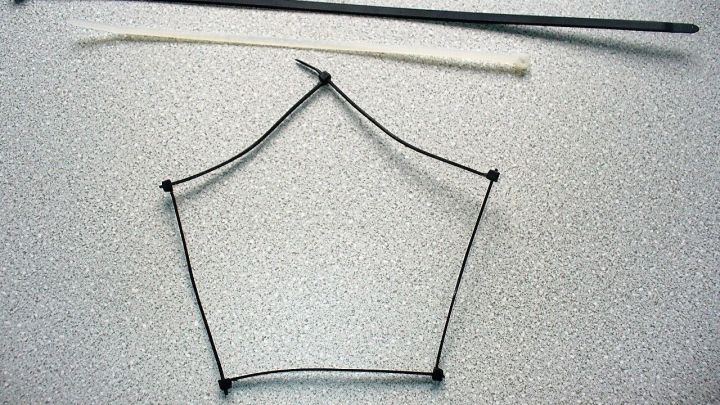 Ein Kabelbinder ist in einem f&uuml;nfeckigen Rahmen angeordnet, w&auml;hrend ein gerader Stab daneben liegt, auf einer hellen Oberfl&auml;che.