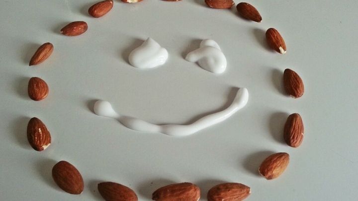 Eine Gesichtscreme aus Mandel&ouml;l liegt in Form eines lachenden Gesichts, umgeben von Mandeln auf einem hellen Hintergrund.