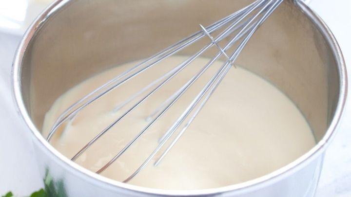 Eine silberne Sch&uuml;ssel enth&auml;lt eine cremige Sauce Hollandaise, w&auml;hrend ein Schneebesen darin r&uuml;hrt. Im Hintergrund sind Butter, Ei, Zitrone und frische Kr&auml;uter zu sehen.