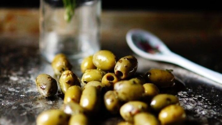 Eingelegte Oliven liegen verstreut auf einer Fl&auml;che, w&auml;hrend eine kleine Blumenvase im Hintergrund f&uuml;r einen rustikalen Touch sorgt.