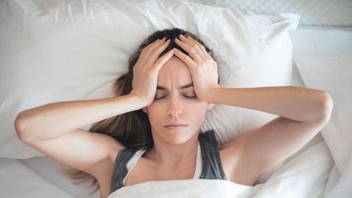 Eine Frau liegt im Bett und h&auml;lt sich mit beiden H&auml;nden den Kopf, um ihre Kopfschmerzen zu lindern.