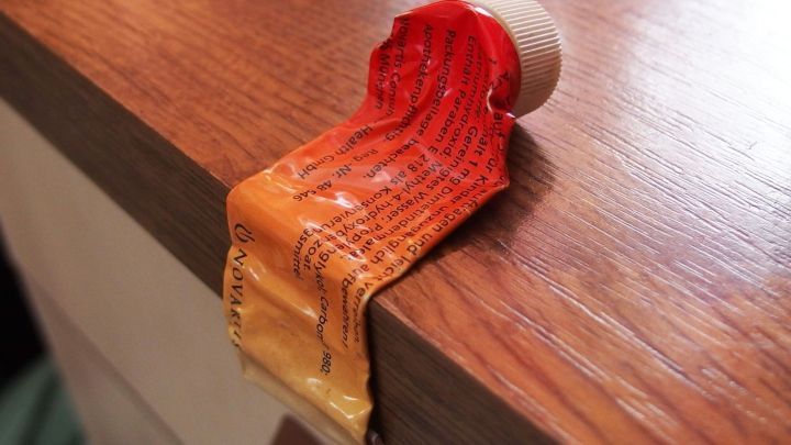 Eine fast leere Tube liegt auf einer Holzoberfl&auml;che, die aufgekr&auml;uselte Tube zeigt Farbreste und ist am Rand des Tisches positioniert.