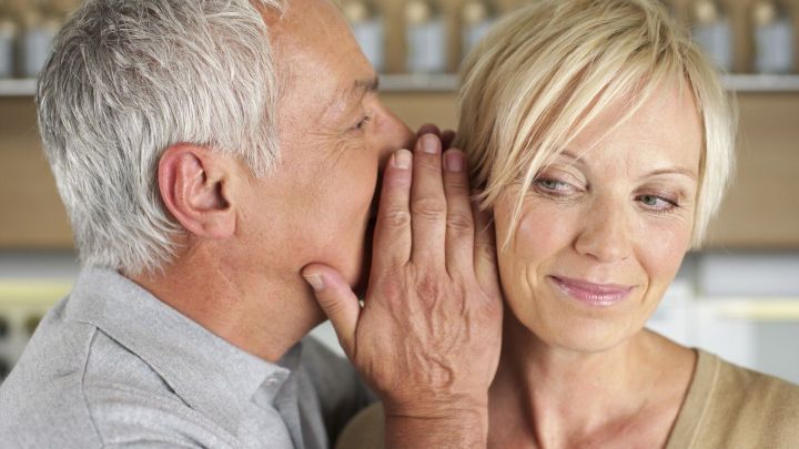 Ein Mann fl&uuml;stert einer l&auml;chelnden Frau ein Kompliment ins Ohr, w&auml;hrend eine freundliche Atmosph&auml;re im Hintergrund herrscht.