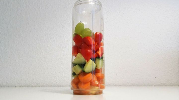 Ein transparenter Smoothie Mixer ist gef&uuml;llt mit bunten Fr&uuml;chten wie Melonen, Trauben und Gurken, vor einem neutralen Hintergrund.